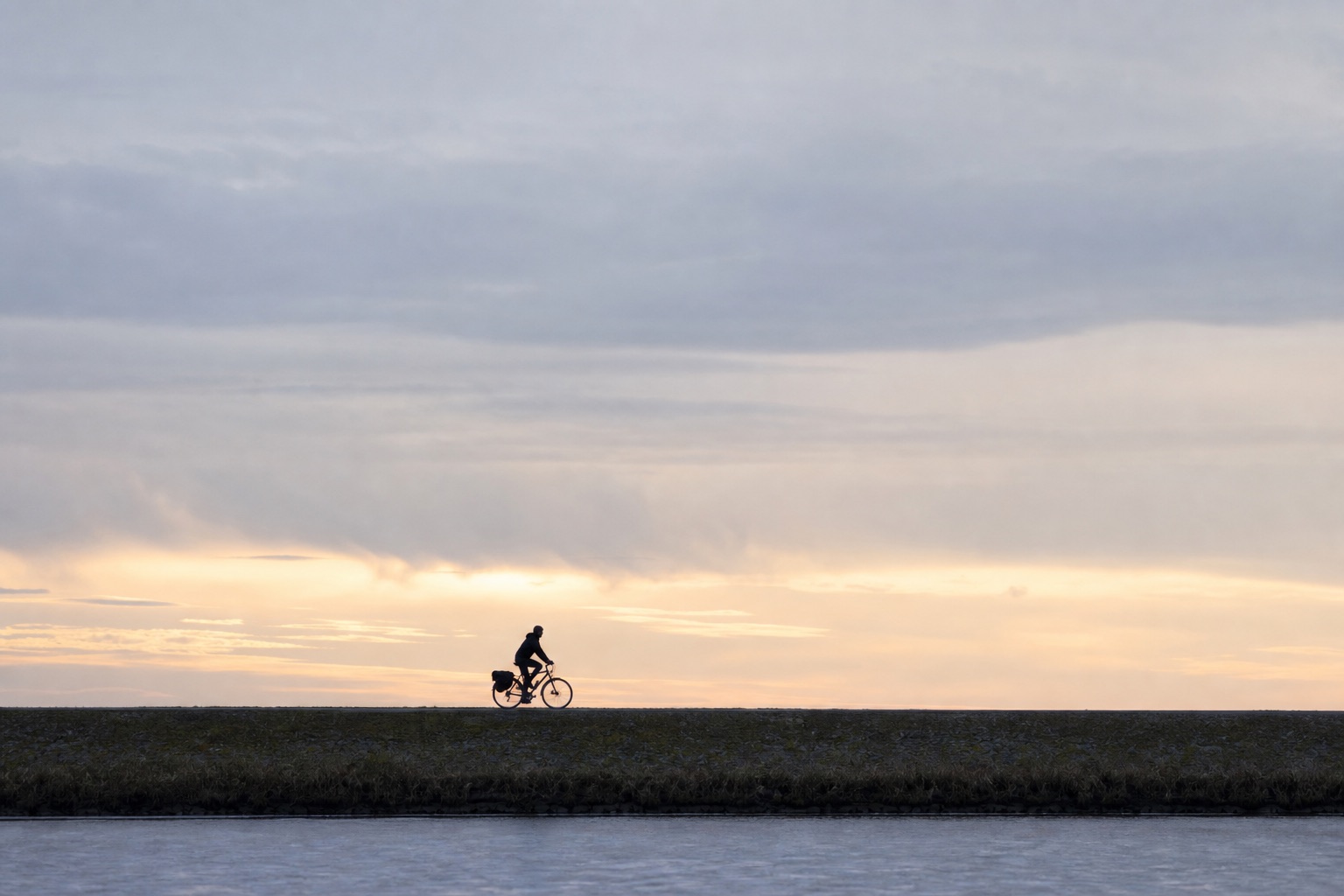 Slim silhouette of a dawn rider on a towpath dyke, grey-blue sky turning pale yellow, minimalist framing